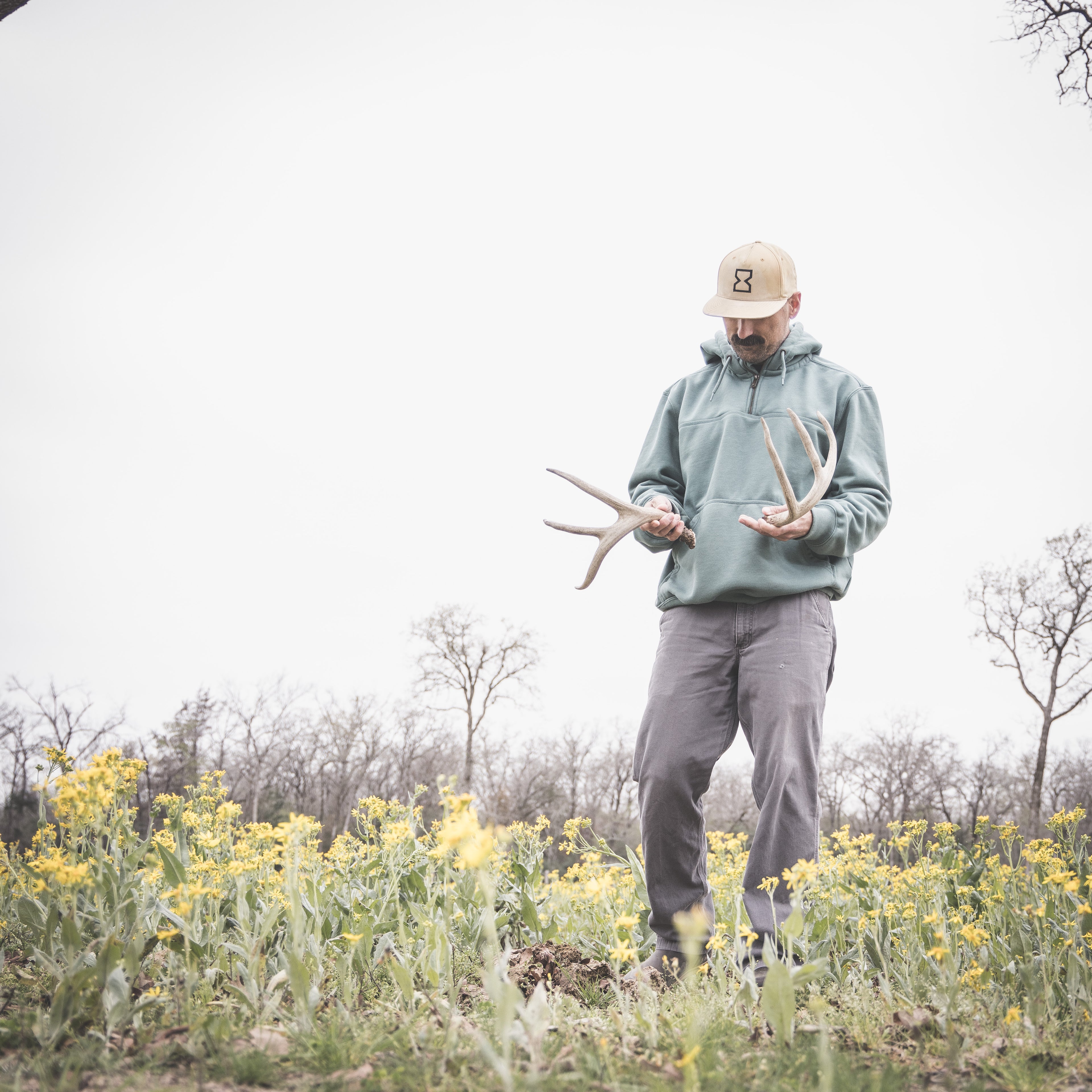 Person holding deer antlers in a field with yellow flowers and bare trees in the background