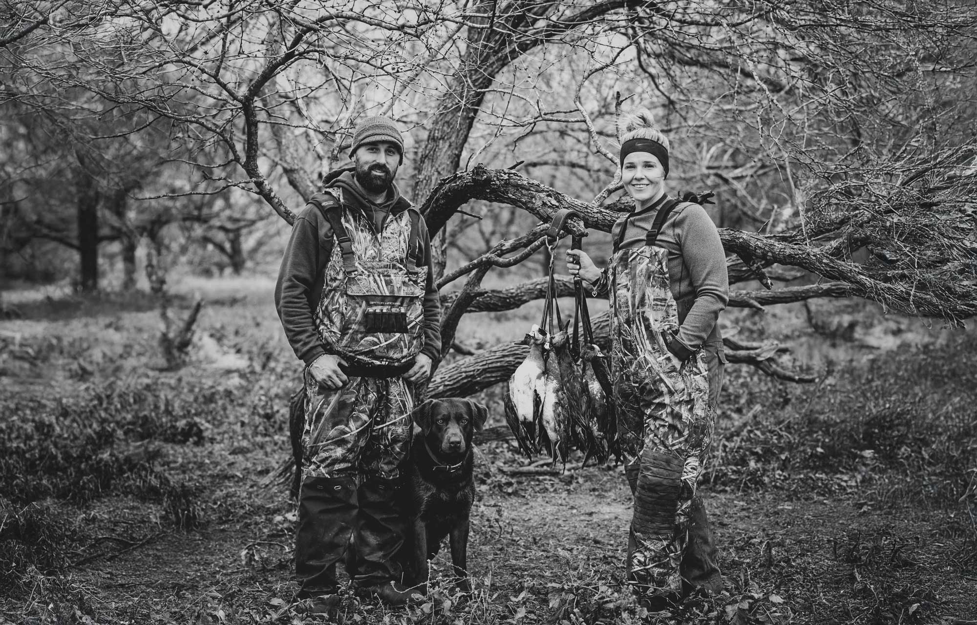 man, woman and dog posing in woods with ducks they caught while hunting