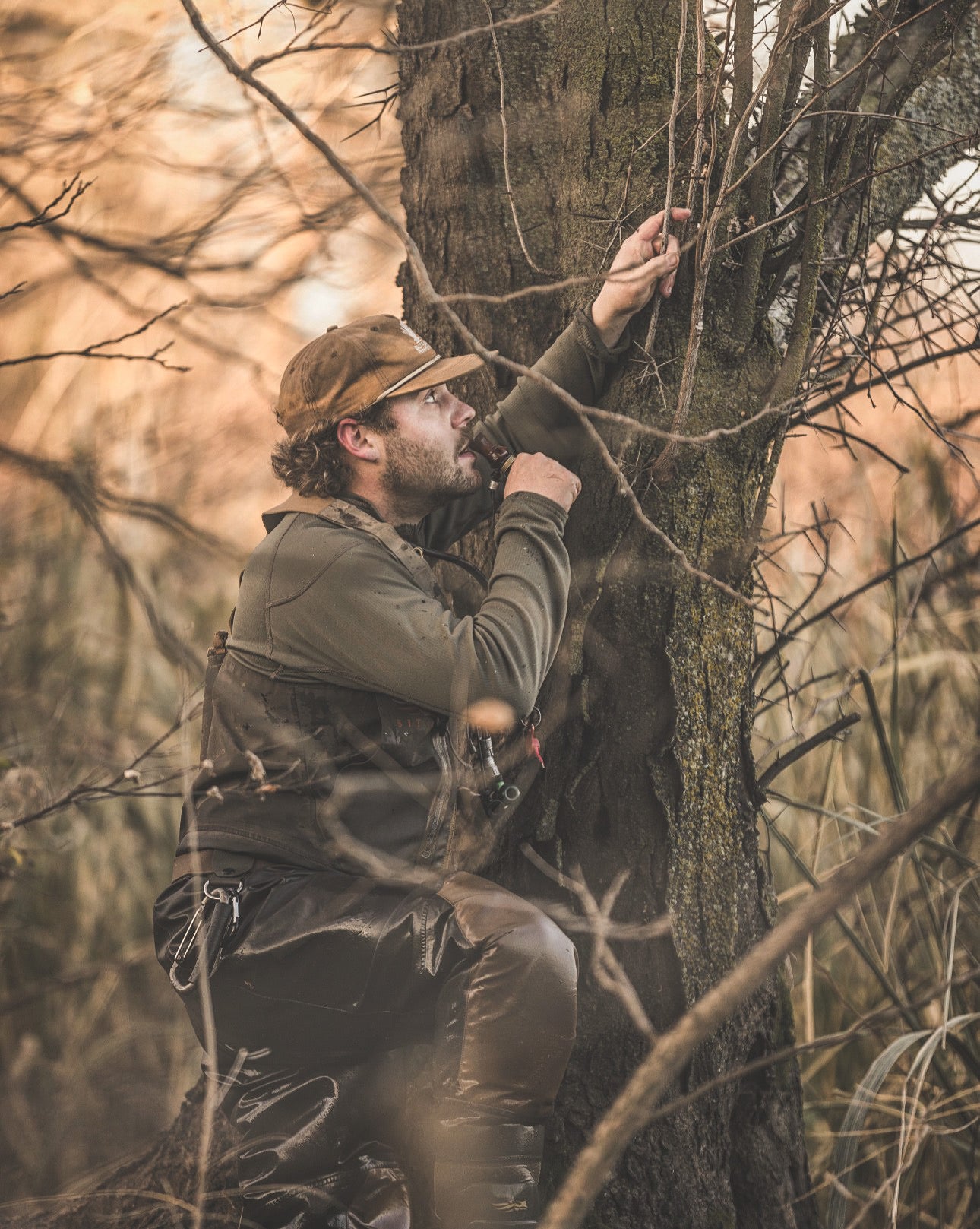 man holding a duck call to his lips as he leans against a tree in the woods