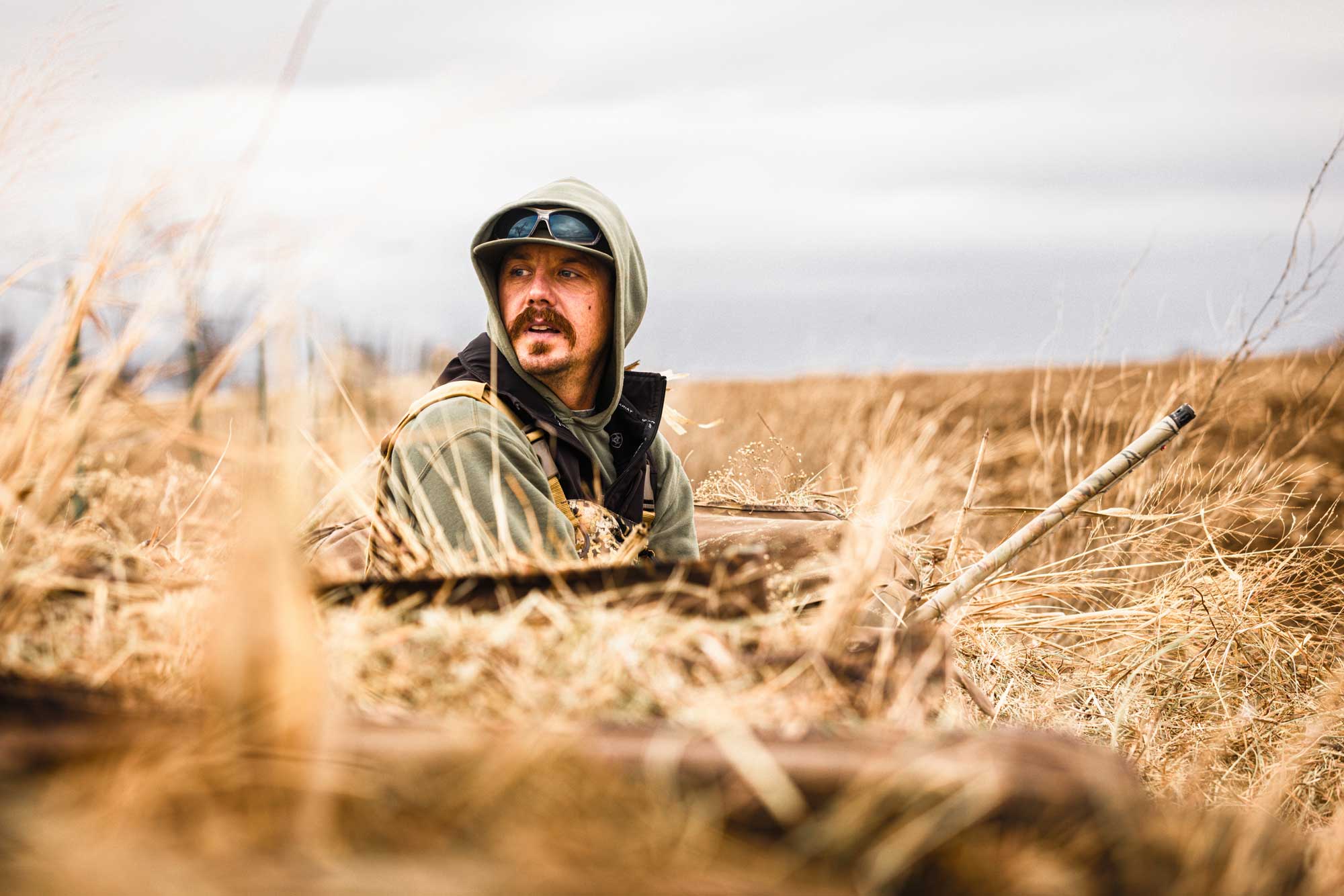 man wearing hunting gear sits in a field and looks off into the distance behind him