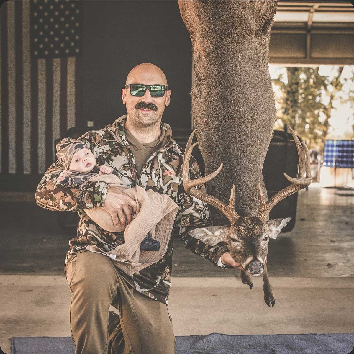 Man holding baby next to hanging trophy deer