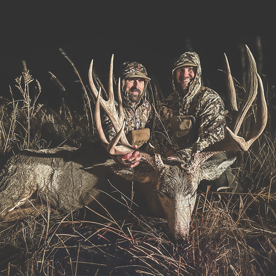two hunters in camo sitting behind a large mule deer, holding up his  9-point antlers